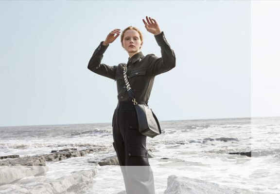 femeie cu camasa kaki, pantaloni negrii si geanta crossbody pe malul oceanului